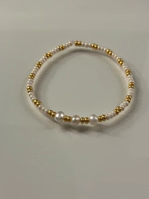 Pärlarmband med vita och guldfärgade detaljer - Snyggt armband med små vita pärlor och guldfärgade metallpärlor i ett stilrent mönster. Armbandet är elastiskt och passar de flesta handleder. Perfekt för dig som gillar en enkel men elegant accessoar.