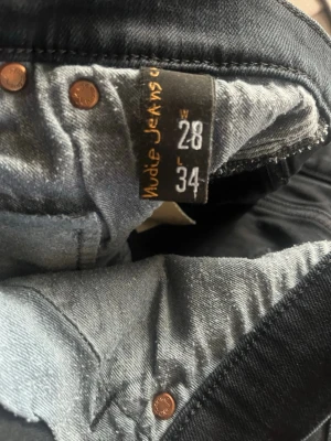 Svarta jeans från Nudie Jeans Co - Säljer ett par svarta jeans från Nudie Jeans Co i storlek 28/34. Jeansen har klassisk femficksdesign, smal passform och är tillverkade i denim med snygg finish. Perfekta för dig som gillar stilrena och tidlösa jeans.