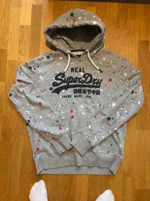 Grå Superdry hoodie med färgstänk - Mått: längd 71cm. Bredd: 65cm. Armlängd 59cm. Skicka gärna bud säljer en fet hoodie. 
