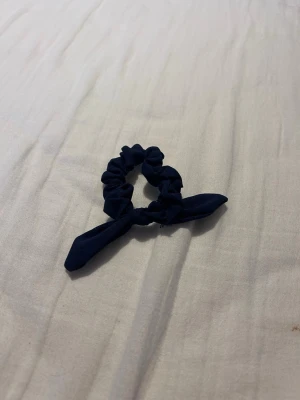 Mörkblå scrunchie med rosett - En stilren mörkblå scrunchie med en söt rosettdetalj. Tillverkad i mjukt tyg som är skonsamt mot håret och ger en trendig look. Perfekt för att piffa upp din frisyr och hålla håret på plats.