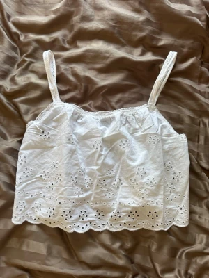 Vit broderad somrig crop top - Söt vit crop top med broderade blommor och hålmönster. Toppen har tunna axelband och en vågig kant nertill. Perfekt för varma dagar och ger en romantisk vibe. Tillverkad i lätt bomullstyg som känns luftigt och bekvämt.