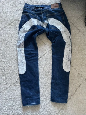 Evisu jeans - Unika mörkblå jeans med en stor vit grafisk målning på baksidan av benen. Klassisk femficksmodell med raka ben och knappgylf. Jeansen har en cool streetwear-vibe och är tillverkade i kraftigt denimtyg.