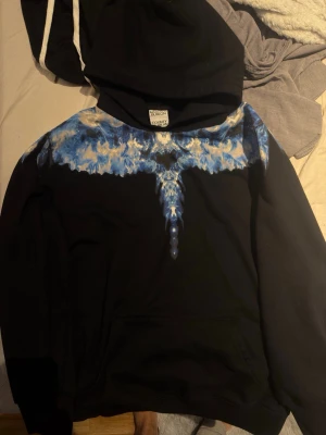 Svart hoodie Marcelo Burlon blått mönster - Säljer en svart hoodie från Marcelo Burlon County of Milan med ett fett blått och vitt grafiskt mönster över axlarna och bröstet. Klassisk huva och känguruficka framtill. Perfekt för dig som vill sticka ut med streetwear vibes.
