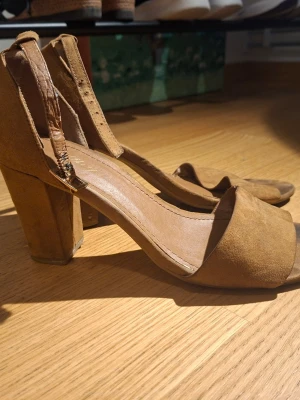 Bruna mocka-pumps med bred klack - Säljer ett par bruna pumps i mockaimitation med bred klack och öppen tå. Skorna har en justerbar rem med spänne runt ankeln och en stilren, minimalistisk design. Perfekta för dig som gillar klassiska men trendiga skor.