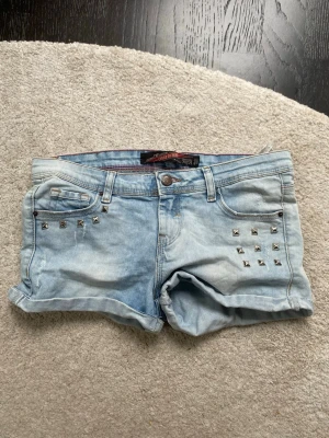 Ljusblå jeansshorts - Snygga ljusblå jeansshorts med upprullade benslut och coola silverfärgade nitar på fram- och bakfickor. Klassisk femficksmodell med bälteshällor och knappstängning. Perfekta för sommaren och ger en edgy vibe till din outfit.