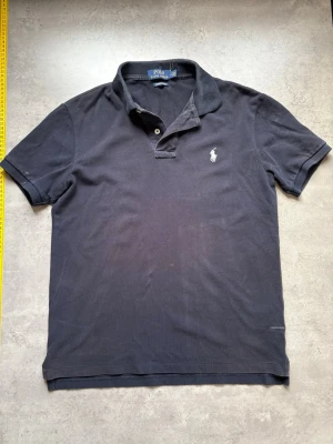 Svart pikétröja från Polo Ralph Lauren - Klassisk svart pikétröja från Polo Ralph Lauren med vit broderad logga på bröstet. Tröjan har korta ärmar, krage och två knappar vid halsen. Tillverkad i bomull för en bekväm och tidlös look.