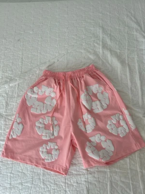Rosa Denim Tear Shorts - Säljer ett par ljusrosa shorts med elastisk midja och stora vita blommönster över hela plagget. Shortsen har en avslappnad passform och är perfekta för varma dagar. Materialet känns mjukt och bekvämt, troligen bomull. Snygg och lekfull design!