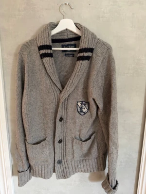 Grå stickad cardigan East West - Grå stickad Cardigan med sjalkrage och svarta ränder på kragen. Koftan har två framfickor, ribbade muddar och knäppning med fyra knappar. På bröstet finns ett broderat emblem i blått och vitt. Perfekt för lager på lager och en avslappnad stil. Knappen längst ned har sytts på i efterhand och är inte den originella men den är snygg ändå och det märks knappast. Skick 7/10, Storlek M, Hör av dig vid frågor🙌