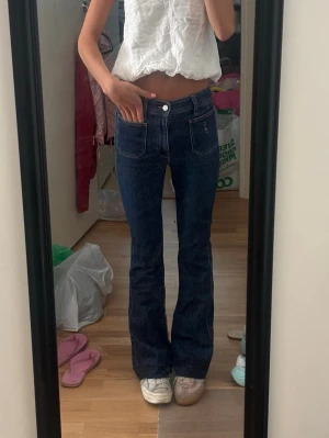 Mörkblå bootcut jeans med fickor - Säljer ett par mörkblå bootcut jeans med hög midja och stora framfickor. Jeansen har en klassisk femficksdesign och är tillverkade i stretchigt denimtyg för en bekväm passform. Perfekta för dig som gillar retrovibbar och vill ha ett par snygga jeans med utsvängda ben. Sprättade längst ned,jag är ca 172 och de passar bra i längden 