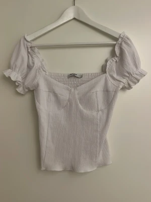 Vit puffärm topp med smock - Supersöt vit topp med puffärmar och smockad rygg. Toppen har en feminin, figurnära passform och korta ärmar med volangkant. Perfekt till jeans eller kjol för en trendig look.