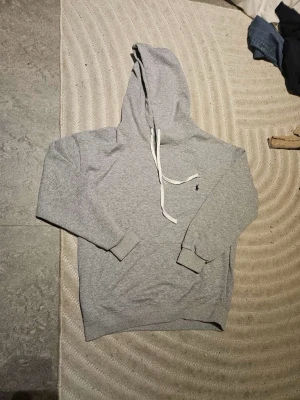 Grå Polo Ralphlauren hoodie  - Klassisk grå hoodie med luva och vita dragsnören. Hoodien har ribbade muddar vid ärmslut och nederkant samt en liten broderad logga på bröstet. Tillverkad i mjukt sweatshirtmaterial som är perfekt för chill dagar.