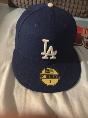 Blå LA 59FIFTY keps från New Era - Snygg blå keps från New Era 59FIFTY med LA-logga broderad framtill i vitt. Klassisk flat peak och strukturerad passform. Perfekt för dig som gillar streetwear och sportig stil.
