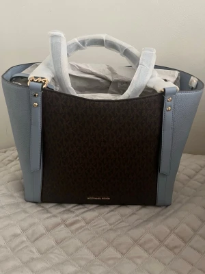 Blå och brun handväska Michael Kors - Snygg handväska från Michael Kors i blått och brunt. Väskan har guldfärgade detaljer, två handtag och är tillverkad i en kombination av slätt och mönstrat konstläder. Perfekt för dig som vill ha en trendig och rymlig väska med exklusiv känsla.