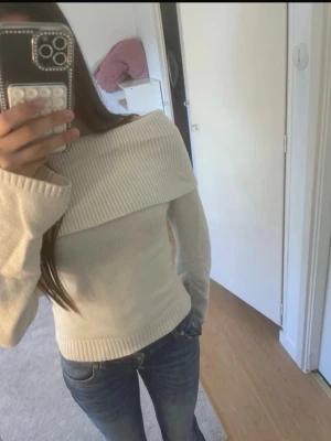 Off shoulder stickad tröja i vitt - Snygg off shoulder stickad tröja i vitt med bred ribbad kant upptill och vid ärmslut. Tröjan har lång ärm och sitter figurnära, perfekt till jeans eller kjol. Materialet är mjukt och stretchigt för en bekväm känsla.