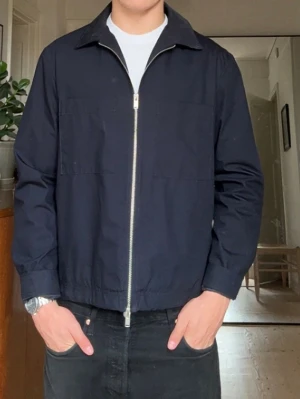 Mörkblå vindjacka med dragkedja - Stilren mörkblå vindjacka med hel dragkedja framtill och klassisk krage. Jackan har raka ärmar och två diskreta bröstfickor. Perfekt för dig som gillar enkel och clean stil.