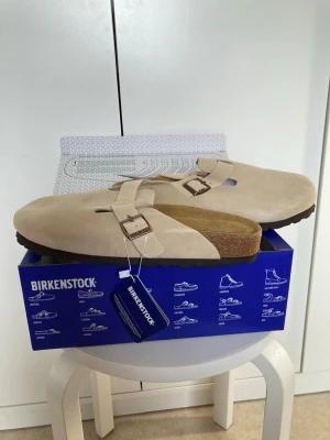 Beige Birkenstock tofflor mocka - Snygga beige tofflor från Birkenstock i mjuk mocka med klassisk design och justerbart spänne i metall. Ergonomisk fotbädd och robust sula för extra komfort. Perfekta för dig som vill ha både stil och bekvämlighet.