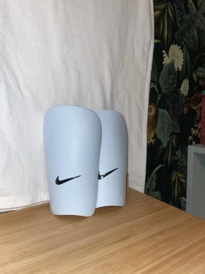 Vita benskydd från Nike - Säljer mina vita nike benskydd som jag inte vet storlek på men skulle chansa på M inte använda så mycket och får med kardborreband och liten påse. Skriv om ni har någon fråga