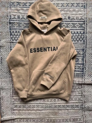 Beige Essentials hoodie med tryck - Helt ny Essentials hoodie, lite för stor på mig därför säljer jag den, 100 procent äkta. Det är en pytteliten fläck på vänstra sidan som ja inte vet vart den kom ifrån, den går säkert bort om man tvättar den. Kom med frågor och bud.  Längd 65 cm Axelbredd ungefär 55 Bredd ner 48 cm