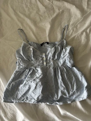 Blåvit randig linne topp från brandy melville med knappar - Söt blåvit randig linnetopp med smala axelband, knappar framtill och knytband i midjan. Toppen har en luftig passform och resår i ryggen för extra komfort. Perfekt och enkel att styla med jeans eller kjol.