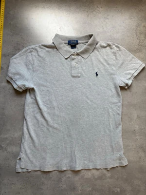 Ljusgrå pikétröja från Ralph Lauren - Klassisk ljusgrå pikétröja från Ralph Lauren med korta ärmar, krage och två knappar framtill. Tröjan har den ikoniska broderade loggan på bröstet och är tillverkad i mjuk bomull. Perfekt för en clean och stilren look.