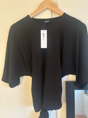 Svart knyt topp från Gina Tricot - En svart topp från Gina Tricot med lös passform och knyt i nederkanten. Toppen har rund halsringning och korta ärmar, perfekt för en chill och stilren look. Tillverkad i mjuk bomull som känns skön mot huden.