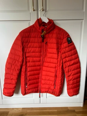 Röd jacka Parajumpers - Röd/orange jacka från Parajumpers med quiltad design, hög krage och gul detalj vid halsen. Jackan har två sidofickor med dragkedja, svart patch med logga på ärmen och ljusgrå insida. Material polyester. Perfekt för kalla vinterdagar.