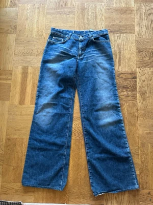 Blåa wide leg jeans med rå kant - Säljer ett par blåa jeans som är Lågmidjade och baggy. Jeansen har klassisk femficksmodell och snygga slitningar framtill och baktill för en avslappnad look. Perfekta för dig som gillar en loose fit och vintagevibe.