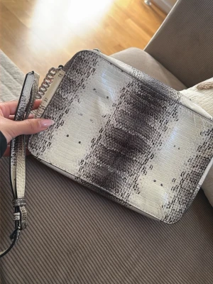 Michael Kors kuvertväska ormmönstrad - Snygg kuvertväska från Michael Kors i ormmönstrat konstläder. Väskan har en stilren svartvit design med silverdetaljer och avtagbar handledsrem. Perfekt accessoar för att lyfta din outfit med en trendig touch.