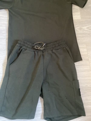 Stone Island set - Snyggt olivgrönt set från Stone Island med t-shirt och matchande shorts. T-shirten har en ikonisk patch på ärmen och rund hals. Shortsen har elastisk midja med dragsko, sidofickor och en praktisk benficka. Perfekt för en avslappnad och trendig look.