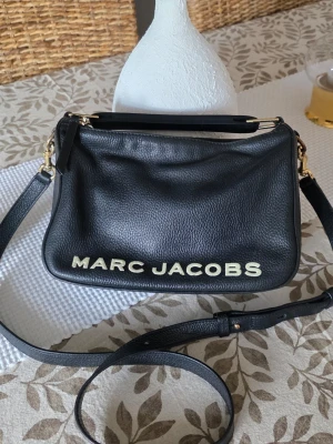 Svart axelväska Marc Jacobs Soft Box 🌿  - Snygg svart axelväska från Marc Jacobs i mjukt läder med justerbar axelrem och guldfärgade detaljer. Väskan har ett stort logotyptryck framtill och en enkel, stilren design som passar till det mesta. Perfekt för dig som gillar trendiga accessoarer. Nypris 5300 kr