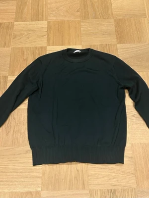 Mörkgrön merinoulltröja från Asket - En mörkgrön stickad tröja från Asket i mjuk merinoull. Tröjan har rund halsringning, långa ärmar och ribbade muddar vid ärmslut och nederkant. Perfekt för lager-på-lager och enkel att matcha med olika outfits.