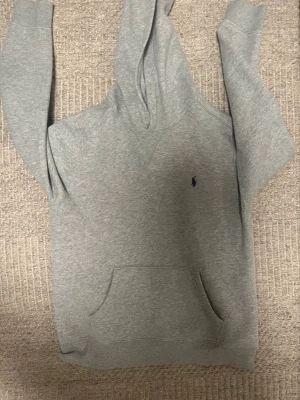 Grå Ralph lauren hoodie - Snygg grå hoodie med huva och känguruficka framtill. Tröjan har en liten broderad logga på bröstet och ribbade muddar vid ärmar och nederkant. Perfekt för en avslappnad och stilren look. Storlek l för ungdomar