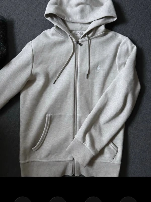 Polo biege hoodie med dragkedja - Snygg biege hoodie med dragkedja och huva. Tröjan har fickor framtill, justerbara snören i huvan och ett diskret broderat logotyp på bröstet. Tillverkad i mjukt sweatshirtmaterial som är skönt mot huden. Perfekt för en avslappnad och stilren look.