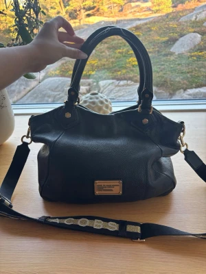 Svart handväska från Marc by Marc Jacobs - Snygg svart handväska i skinn från Marc by Marc Jacobs med guldfärgade detaljer och en bred axelrem med texten 'MARC'. Väskan har dubbla handtag och en metallbricka framtill. Perfekt för dig som vill ha en stilren och rymlig väska. Axelrem bandet var precis bytt så det är helt nytt. Därför säljes den lite dyrare