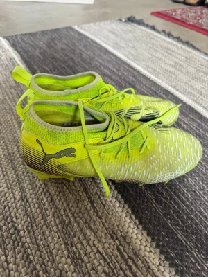 Gula Puma fotbollsskor med snörning  - Säljer ett par snygga fotbollsskor från Puma i neon-gult med svarta detaljer och vita mönster. Skorna har snörning, högre strumpliknande skaft och är tillverkade i syntetmaterial för bästa grepp och känsla på planen.