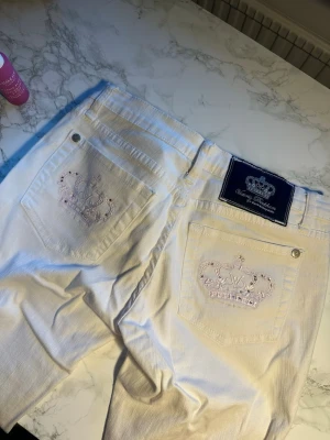 Vita Victoria Beckham jeans  - Säljer ett par vita jeans från Victoria Beckham med broderade kronor och strasstenar på bakfickorna. Klassisk femficksmodell med raka ben och silverfärgade nitar. Snygg detalj med stor blå patch bak i midjan. Perfekta för dig som gillar statement-plagg. Enda defekten är på sista bilden men tycker verkligen inte det syns när de sitter på. De är bootcut. 
