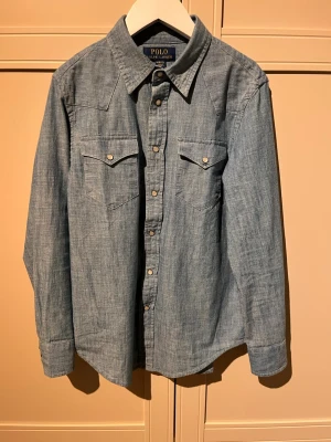 Blå jeansskjorta från Polo Ralph Lauren - Säljer en klassisk blå jeansskjorta från Polo Ralph Lauren. Skjortan har tryckknappar framtill, två bröstfickor med lock och lång ärm. Perfekt för dig som gillar en avslappnad men ändå stilren look.