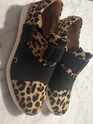 Sneakers med leopardmönster och resår - Säljer ett par snygga sneakers med leopardmönster och breda svarta resårband över foten. Skorna har vit sula och är i en låg modell. Perfekta för dig som vill sticka ut med din stil och gillar djurmönster.