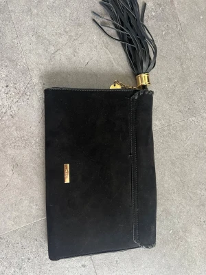 Svart mocka clutch med tofs från Aldo - Snygg svart clutch i mocka med dekorativ guldfärgad detalj och stor tofs. Väskan har en guldfärgad kedja och ett litet metallmärke framtill. Perfekt för dig som vill ha en stilren och trendig accessoar.