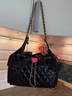 Svart quiltad handväska med kedja - Snygg svart handväska i quiltat konstläder med guldfärgad kedja som axelrem och dragsko. Väskan har en rosa Hello Kitty-detalj framtill och trendig bucket bag-form. Perfekt för dig som gillar en edgy och lekfull stil.