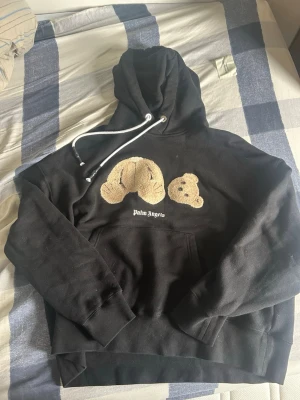 Palm Angels Teddy Bear Hoodie - Svart Palm Angels-hoodie med den ikoniska teddybjörn-grafiken (en björn som håller för ögonen + en mindre björnhuvud). •  100 % bomull •  Broderade teddybjörnar i chenille/terry-look •  Palm Angels-logo under grafiken •  Dragsko i huva med logotyp •  Kanguru-ficka •  Ribbstickade muddar och nederkant •  Storlek: L •  Made in Italy •  Artikelnummer: PMBB058R21FLE001