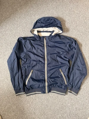 Marinblå vindjacka från H&M L.O.G.G. - Snygg marinblå vindjacka från H&M L.O.G.G. med huva och vita detaljer. Luvan går att ta av, hör av dig vid frågor och behova av fler bilder!!!!!🤗🤗🤗🤗