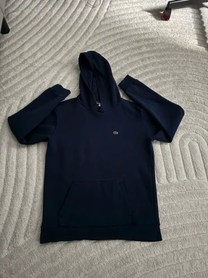 Lacoste Mörkblå hoodie med logga - Lacoste Säljer en mörkblå hoodie med huva och magficka. På bröstet finns en liten broderad logga i form av en krokodil. Hoodien har långa ärmar och ribbade muddar. Perfekt för dig som gillar en clean och stilren look.