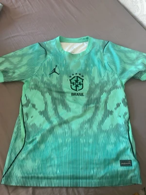 Brasilien  - Säljer en grön träningströja med Brasilien CBF-logga och svart mönster. Tröjan har korta ärmar, rund hals och är tillverkad i ett lätt, snabbtorkande material som passar perfekt till fotbollsträning eller gym. Snygg design med svarta detaljer och modern look.