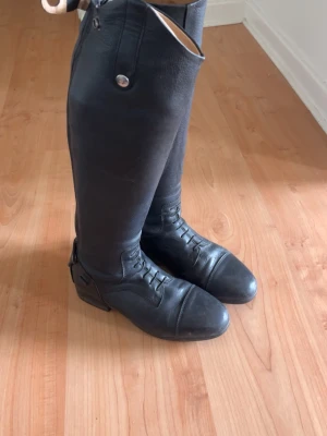 Svarta höga ridstövlar i läder - Säljer ett par svarta höga ridstövlar i läder med snörning framtill och dragkedja bak. Klassisk design med rund tå och dekorativa sömmar. Perfekta för dig som vill ha en stilren och tidlös look till ridningen. Stövelstöd medföljer 