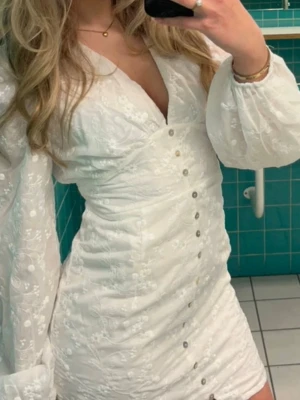 Vit kort klänning med broderi - Säljer en vit kort klänning med v-ringning, långa puffärmar och fina broderade blommor. Klänningen har knappar framtill och markerad midja som ger en snygg siluett. Perfekt för sommarens alla tillfällen!