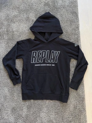 Svart hoodie från Replay med tryck - Svart hoodie från Replay med stor REPLAY-logga och texten 'DENIM GOODS SINCE 1981' på bröstet. Klassisk känguruficka framtill och huva. Långa ärmar med muddar och mjukt material som känns skönt mot huden. Perfekt för en avslappnad streetwear-look.