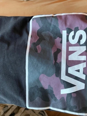 Svart Vans t-shirt med lila camo - Säljer en svart t-shirt från Vans i storlek medium. Framsidan har ett stort tryck med lila och svart camouflage-mönster och tydlig Vans-logga. T-shirten är i 100% bomull och har en klassisk passform. Perfekt för dig som gillar streetwear och vill sticka ut.