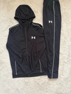 Svart set från Under Armour - Snyggt svart träningsset från Under Armour. Setet består av en hoodie med dragkedja och matchande byxor, båda med logga på bröst och lår. Perfekt för gymmet eller chill. 