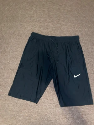 Svarta träningsshorts från Nike - Säljer ett par svarta träningsshorts från Nike med klassisk vit logga på benet. Shortsen har elastisk midja med snörning och två sidofickor. Perfekta för gymmet eller sport. Lätta och bekväma att bära.